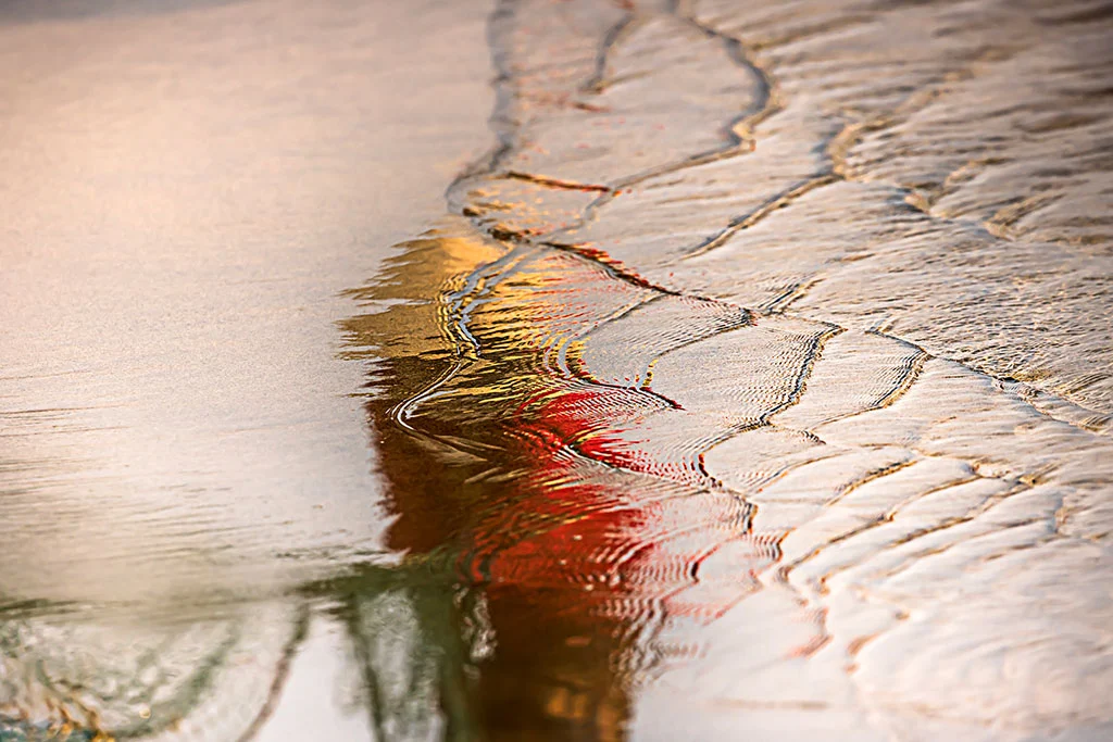 Reflection of a Vietnamese fisherman wearing a conical hat, seen in vivid red and yellow tones through rippling water on a beach in Hoi An, Vietnam.