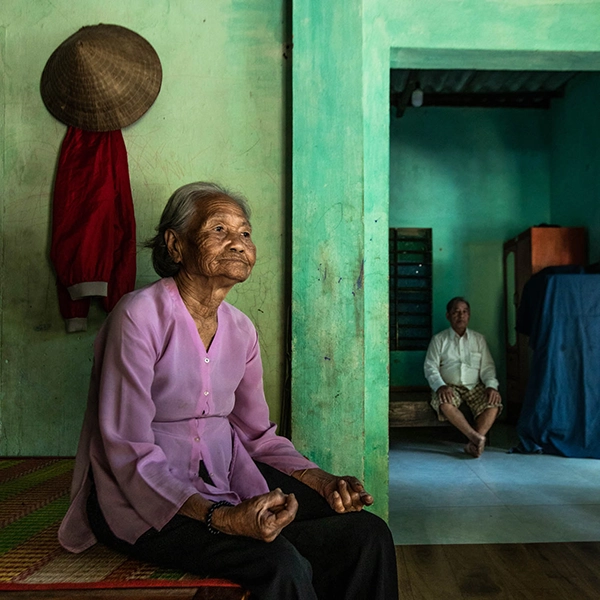 Elderly Vietnamese woman sitting on a mat inside a green house while a man sits in the next room behind her
